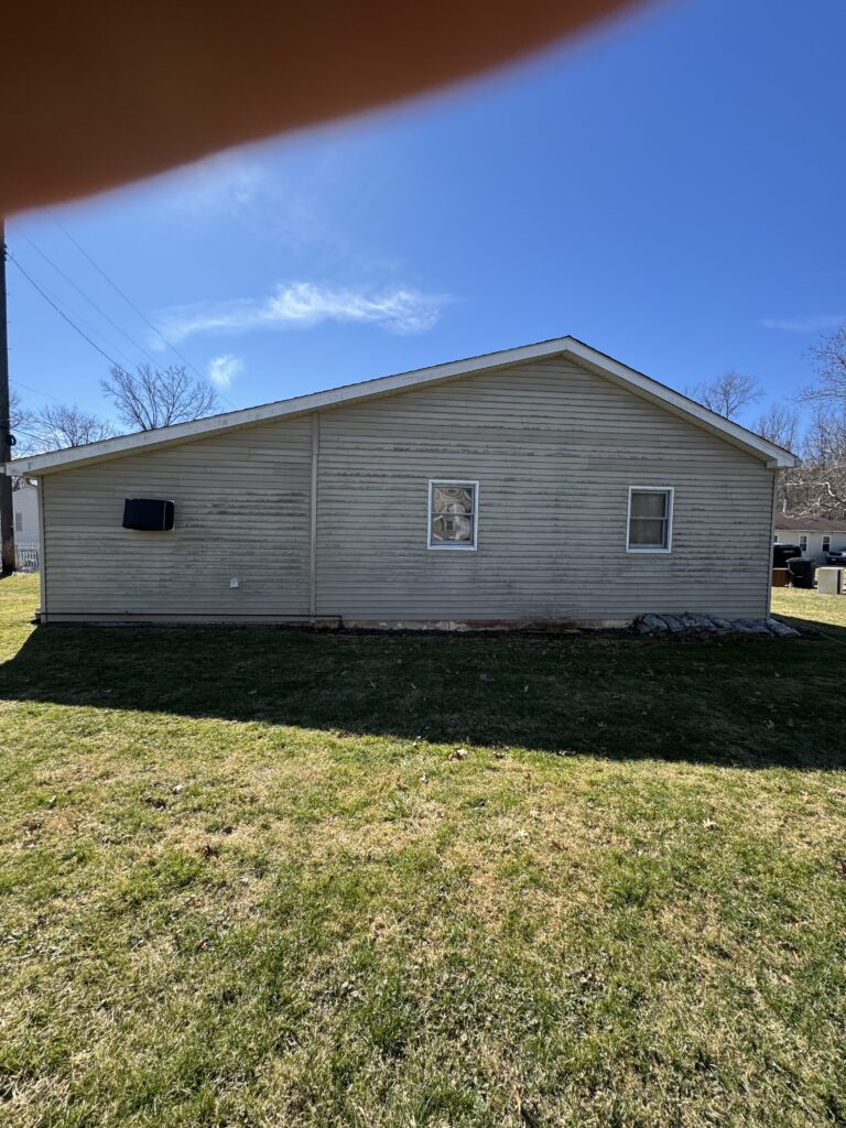 Side view of a beige, single-story house with two windows on a grassy yard under a bright blue sky; a large brown shape in the top-left is a finger covering part of the frame.