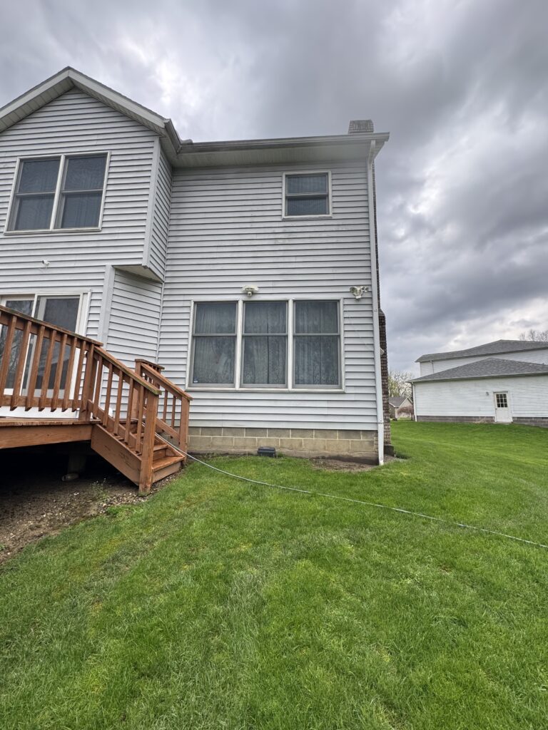 Two-story house with gray siding, large windows, and a wooden staircase railing in the yard under a cloudy sky.