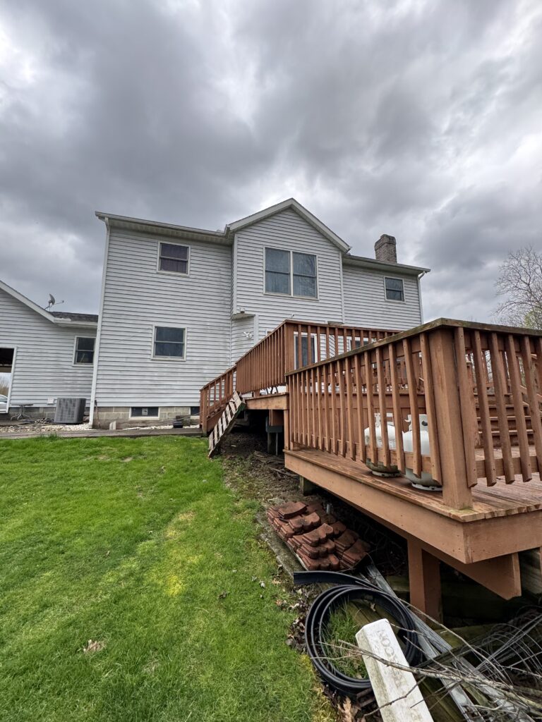 Backyard view of a white two-story house with a wooden deck, stairs, and coiled hoses under a cloudy sky.