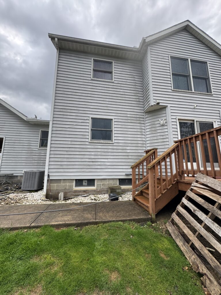 Rear view of a two-story house with gray vinyl siding, a wooden deck staircase on the right, and an outdoor air conditioner unit near the foundation under a cloudy sky.