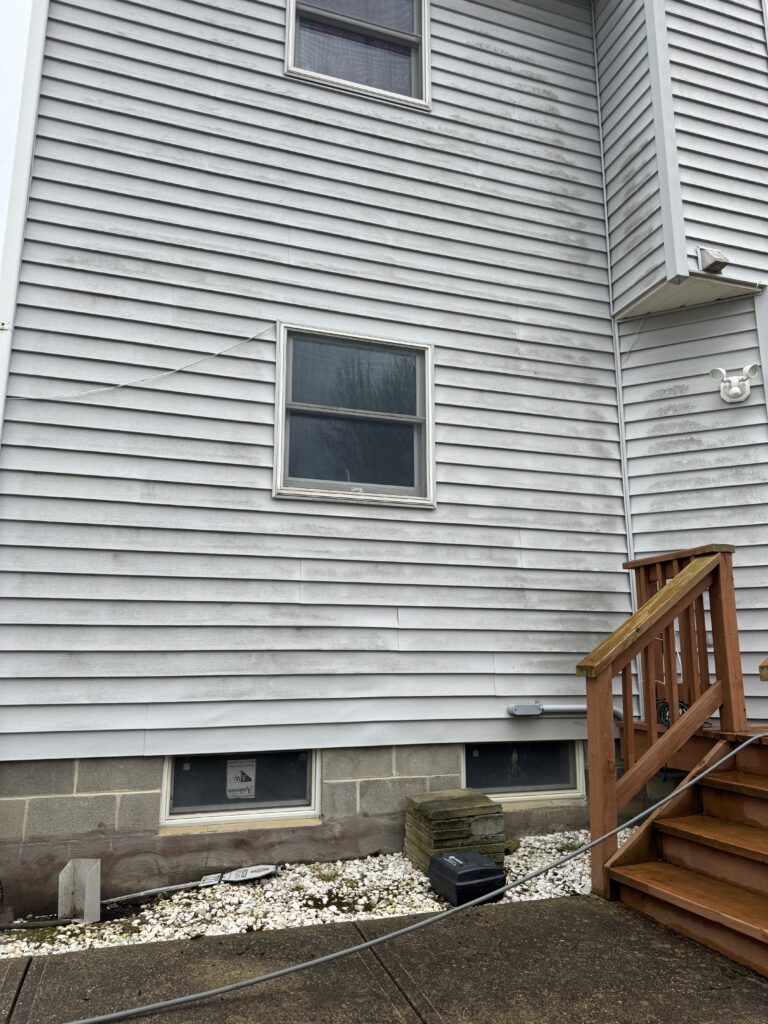 Exterior of a house with light gray horizontal siding, two small basement windows, and a wooden staircase on the right.