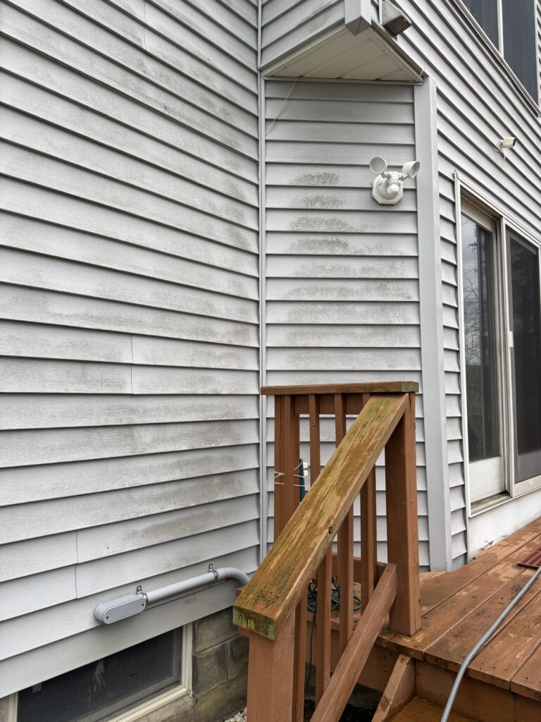 Side of a light-gray, horizontally paneled house with a small wooden deck railing and stairs, showing moss on the railing and siding near a sliding door.