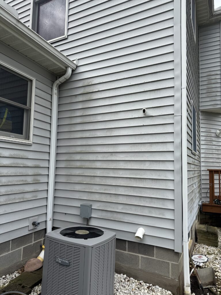 Outdoor air conditioner unit on gravel beside a gray siding house, with white downspouts and a small wall-mounted security camera nearby.