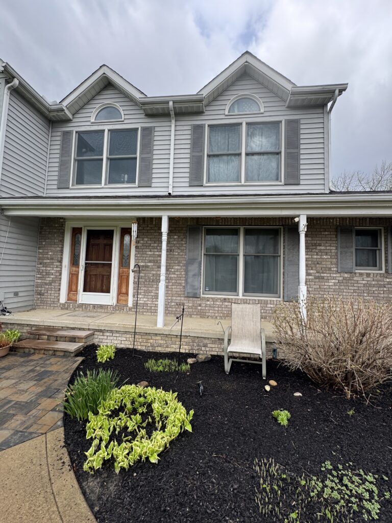Front view of a two-story house with gray siding, brick lower level, and a small garden bed with variegated green plants in front of the porch.