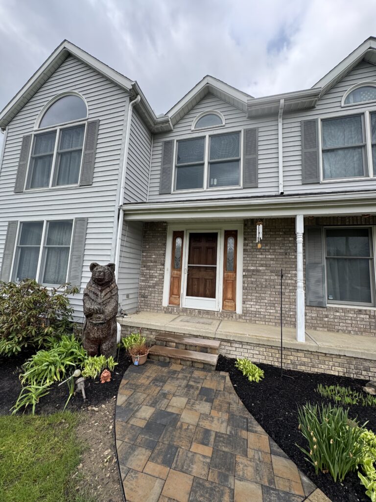Front entrance of a two-story house with gray siding, arched windows, and a brick lower level; a stone bear statue sits near a landscaped garden path.