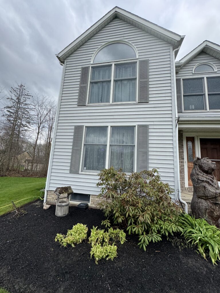 Front view of a two-story suburban house with light blue siding, arched upper window, and gray shutters, a mulched front yard with shrubs and trees in the background.
