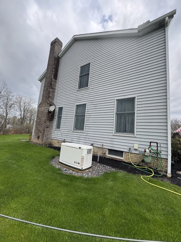 Backyard view of a two-story house with a brick chimney, gray siding, and four windows; lawn and outdoor equipment nearby (AC unit, hoses).