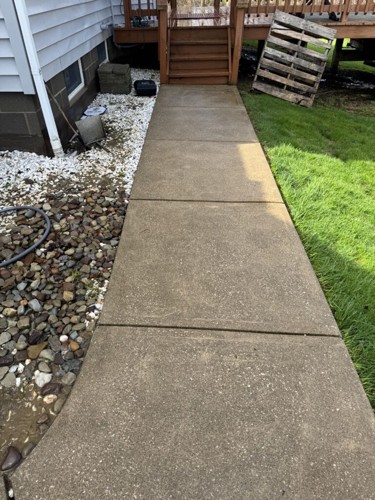 Concrete walkway to a raised wooden deck with stairs; left side has gravel and rocks, right side green lawn, and a leaning wooden pallet nearby.