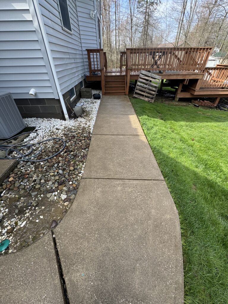 Concrete pathway along the side of a house with a visible long crack, next to a grassy lawn and a wooden multi-level deck.”,