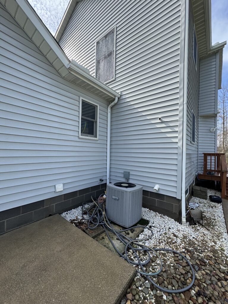 Exterior view of a light gray siding house with an A/C unit on a gravel and stone ground, hoses coiled nearby under a downspout.