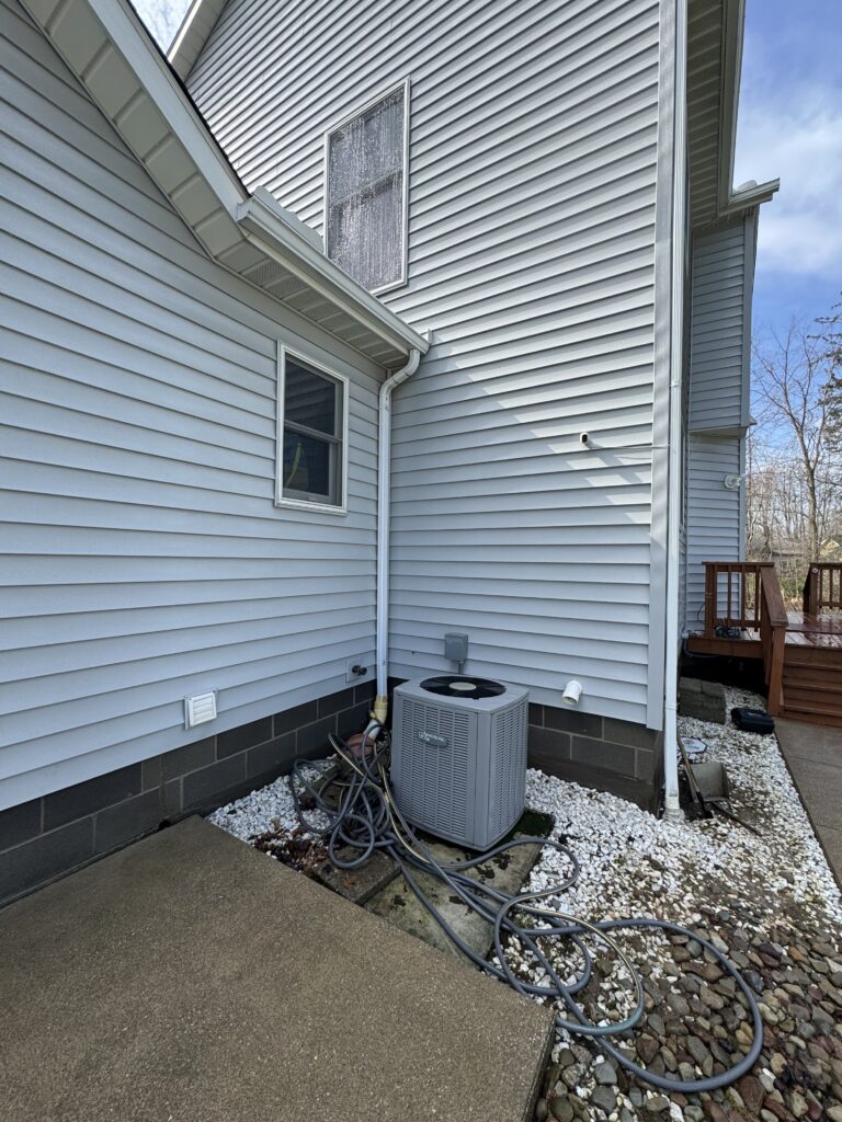 Outdoor air conditioner unit beside a light-blue house, with a tangle of hoses on gravel and white decorative rocks nearby.