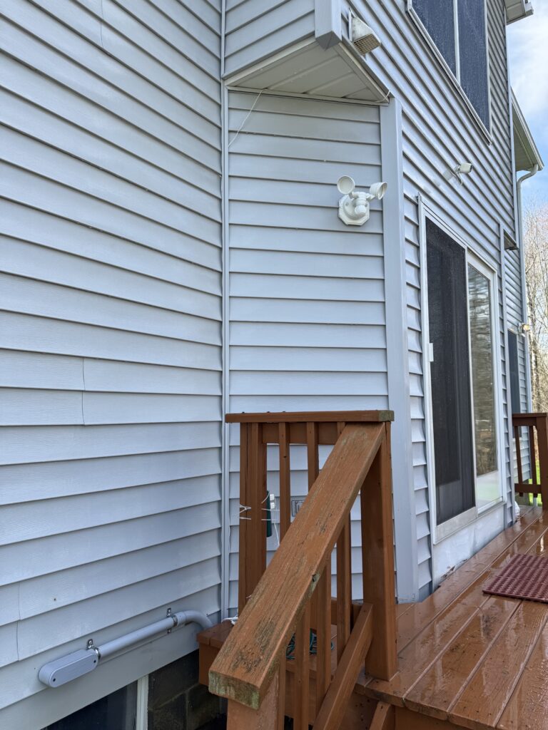 Side of a blue-gray vinyl-siding house with a wooden deck railing, a white exterior light fixture on the wall, and a sliding glass door