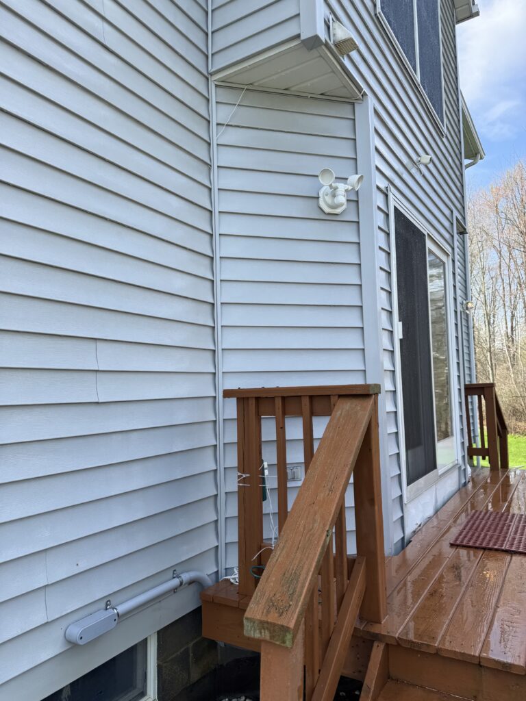 Blue house exterior with vinyl siding, wooden deck stairs, and a white wall-mounted dual-lamp security light above a doorway.