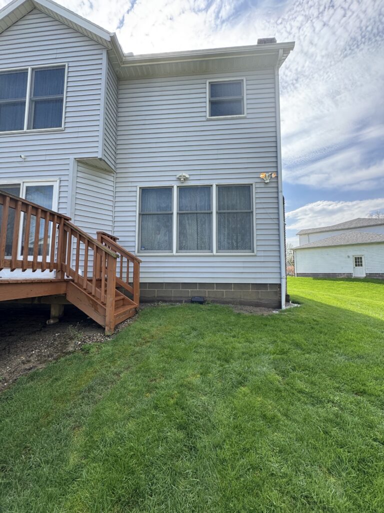 Rear view of a two-story house with gray siding, wooden deck stairs, and large windows overlooking a green lawn under a partly cloudy sky.