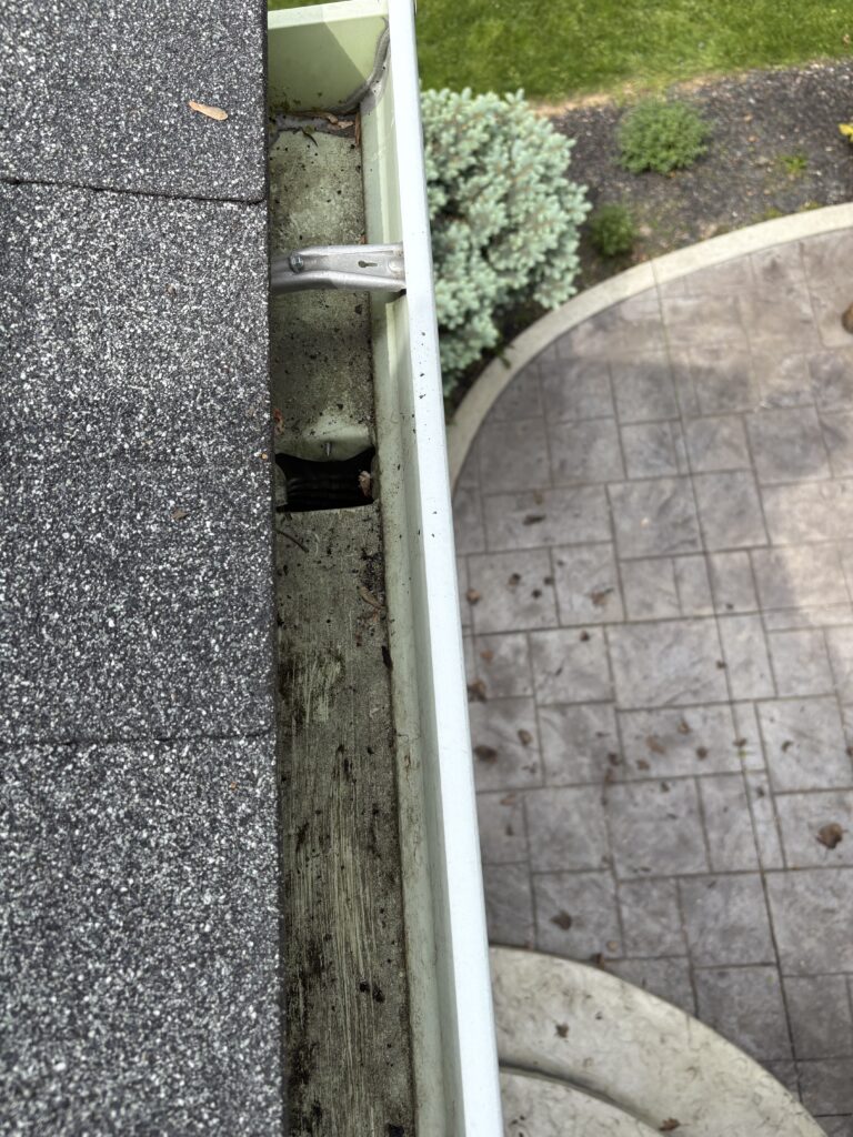 View from the roof edge showing a metal gutter and downspout attached to a weathered wooden fascia, with dark asphalt shingles above and a paved courtyard below.