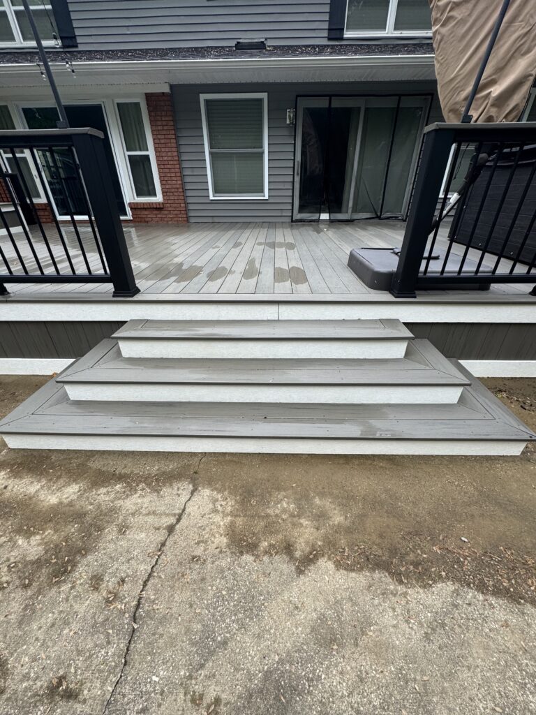 Backyard deck with gray wooden planks, black railing, and a sliding glass door leading into a house; wet spots on the deck surface and steps visible.