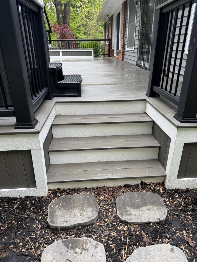 Wooden deck with gray stairs and black railings, wet from rain, leading to a sliding door on a house with gray siding and brick accents.