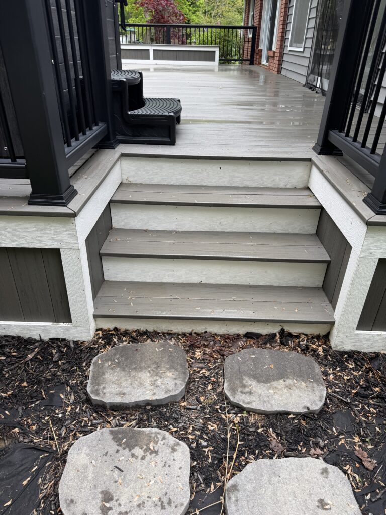 Raised wooden deck with black railing, gray steps, and large flat stepping stones over mulch in a backyard.”