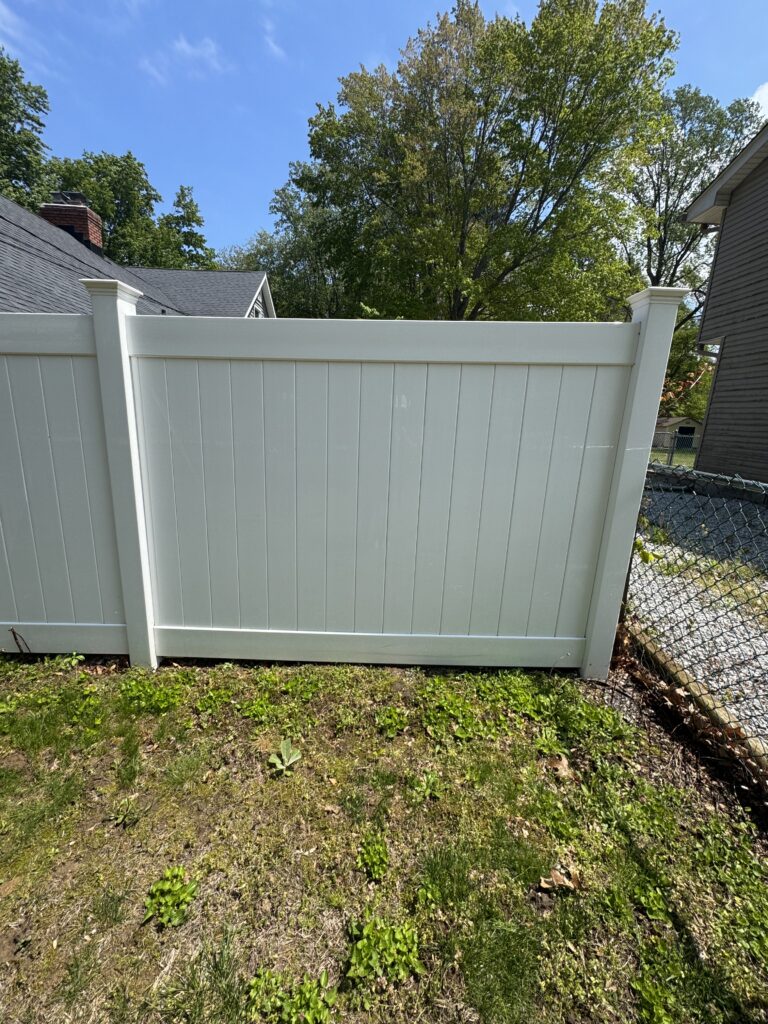 White vinyl privacy fence with vertical panels and capped posts in a backyard, blue sky and green trees in the background.