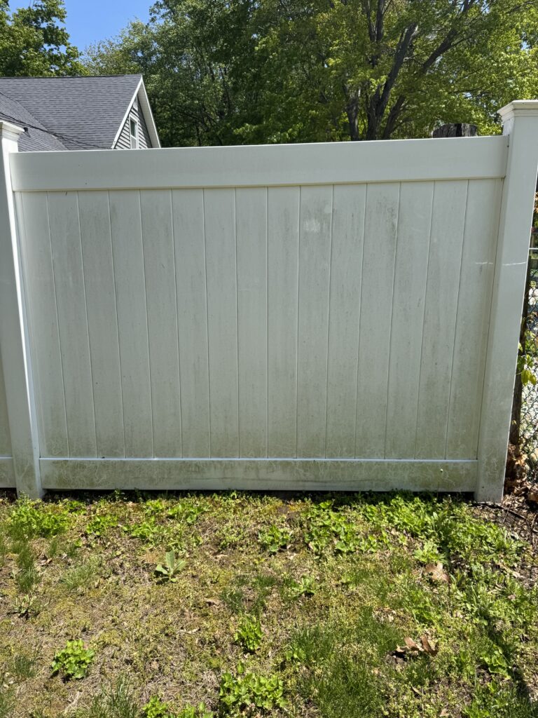 White vinyl privacy fence panel with top rail in a sunlit backyard; a house roof and leafy trees visible in the background.