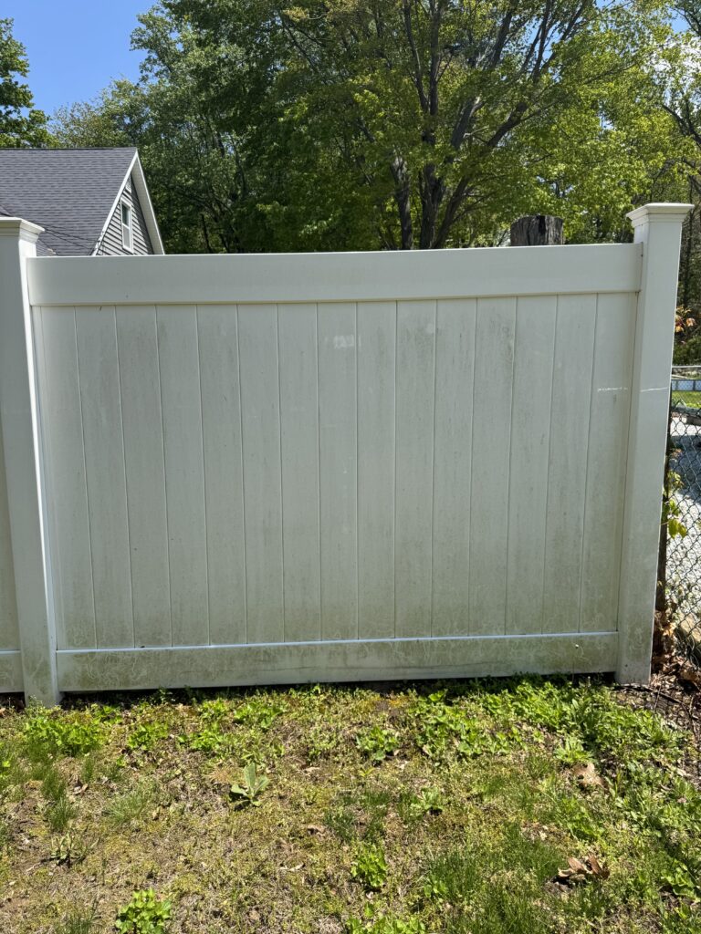 White vinyl privacy fence with vertical panels and posts, set in a yard with grass and overgrown weeds, trees behind it.