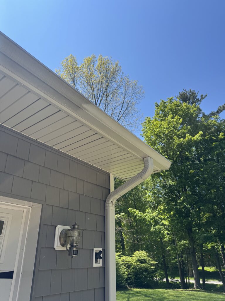 Gray house exterior with white gutter along the roofline, outdoor lantern on the wall, and green trees against a bright blue sky in the background.