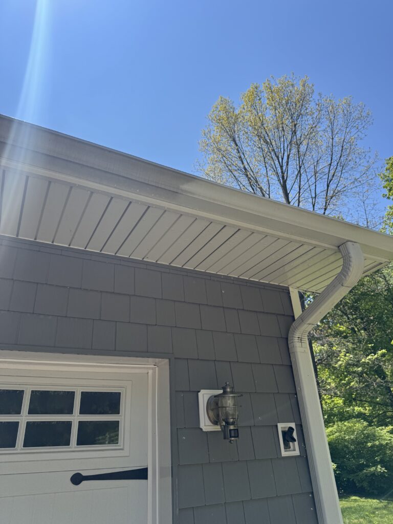 Partial view of a gray house exterior with a white gutter and downspout, a lantern-style light fixture, and a garage door, under a clear blue sky with a tree in the background.