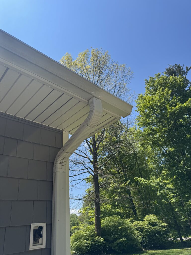 Corner of a gray house with a white gutter and downspout, blue sky and green trees in the background.