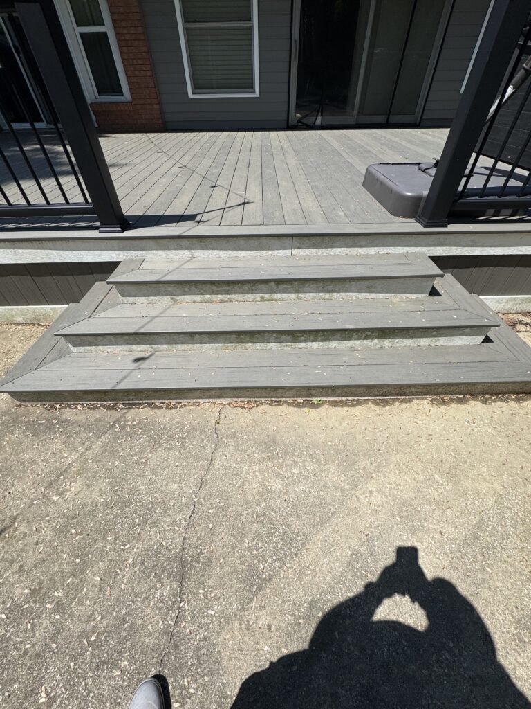 Top-down view of a gray wooden deck with stairs, metal railing posts, and a distant house exterior; photographer's shadow appears in the foreground.