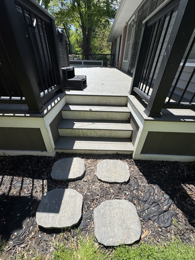 Wide deck with a short flight of stairs and black railing, leading to a yard with stepping stones on a mulch path and green grass nearby.