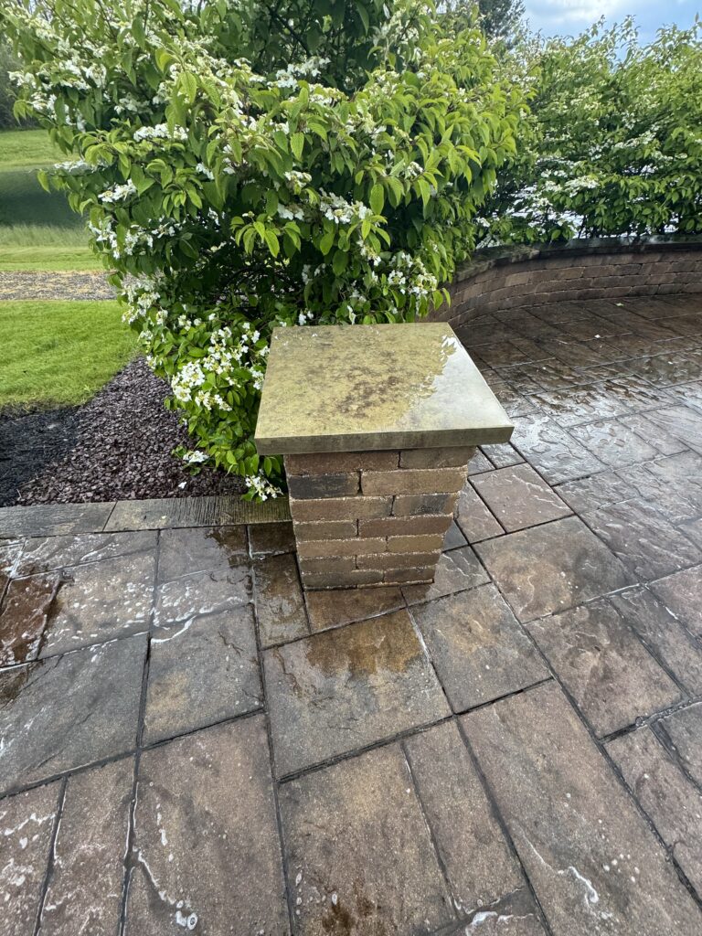 Wet stone square pedestal with water stains beside a flowering shrub with white blossoms on a curved brick wall patio.