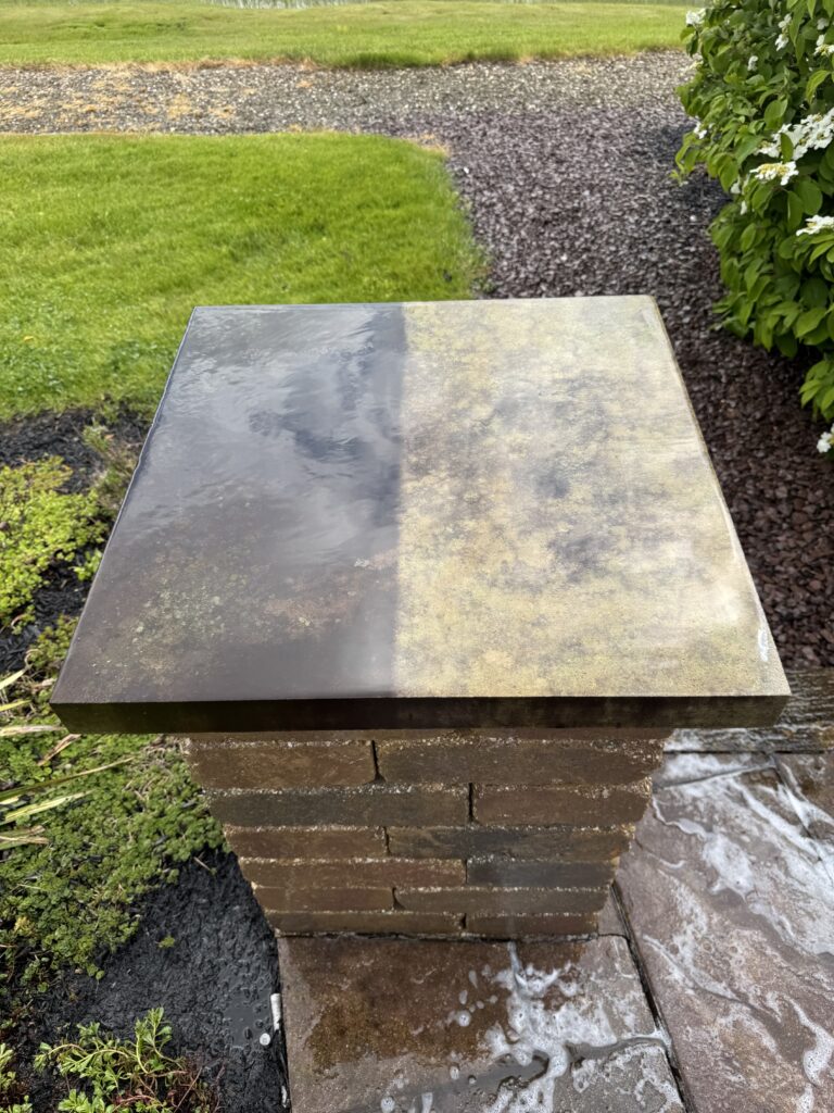 Square, weathered metal tabletop atop a brick pedestal in a garden, with green lawn and shrubbery nearby.