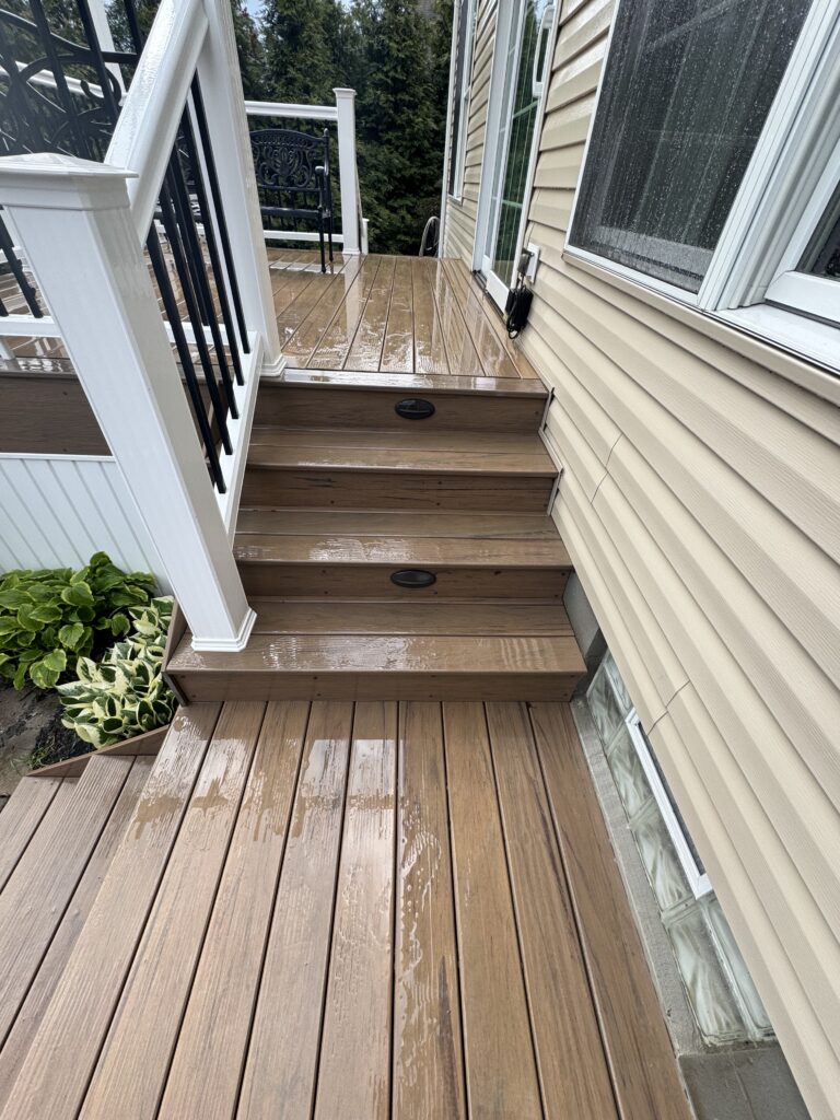 Wet wooden deck and steps outside a beige house, with black metal railing, a sliding door, and green plants in a planter nearby after rain.