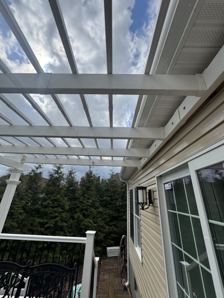 White pergola beams over a back patio attached to a beige siding house, with a sliding glass door and outdoor lamp nearby.