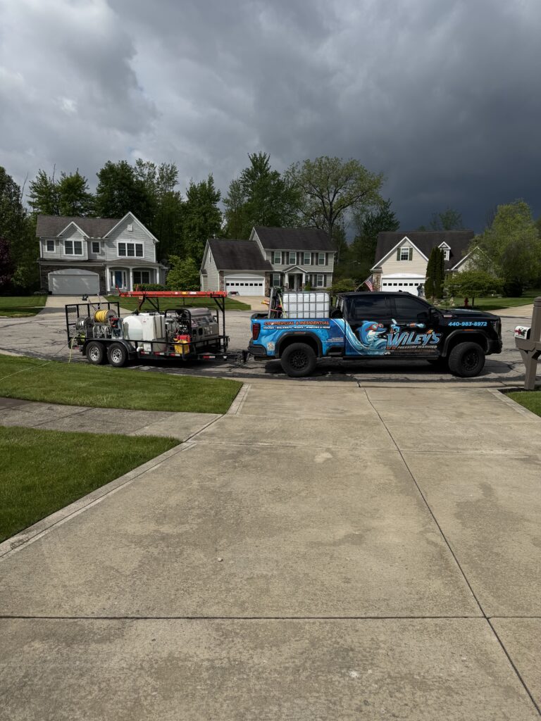 Blue Wiley's pressure washing truck and equipment trailer parked on a suburban street in front of three houses as storm clouds loom.