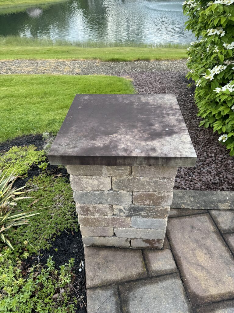 Stone pedestal with a wide flat cap resting on stacked blocks in a landscaped garden by a lawn and lake.