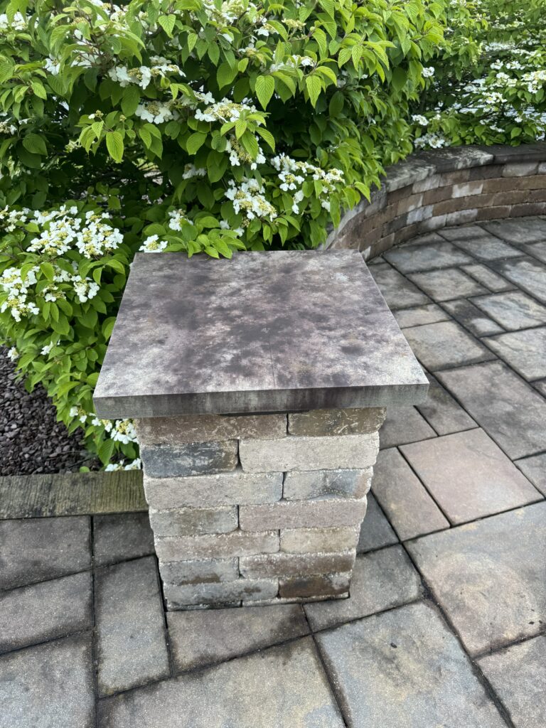 Stone pedestal on a stacked-stone base in a garden, with green shrub and white blossoms behind, and a curved brick wall/paved patio nearby.