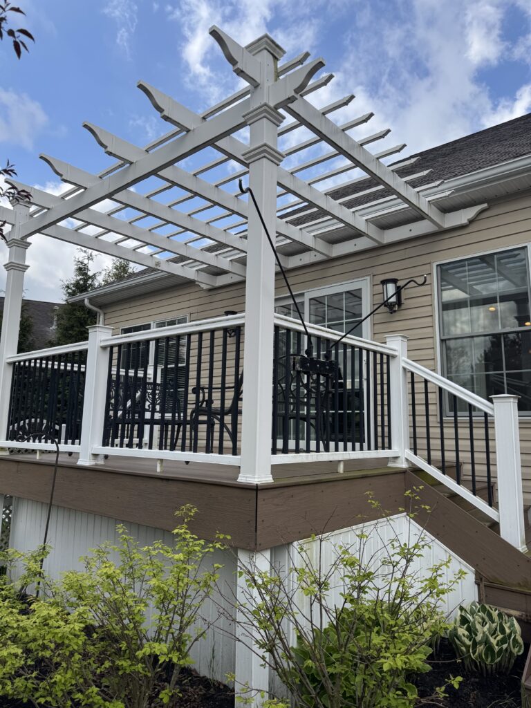 White wooden pergola over a deck beside a beige house, with black railing and green shrubs in the foreground.