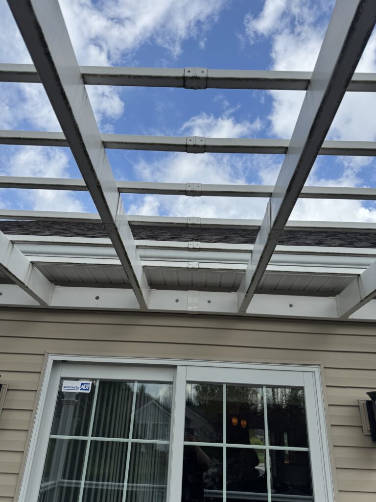 White metal pergola over a sliding glass door on beige siding, with a blue sky and clouds above.