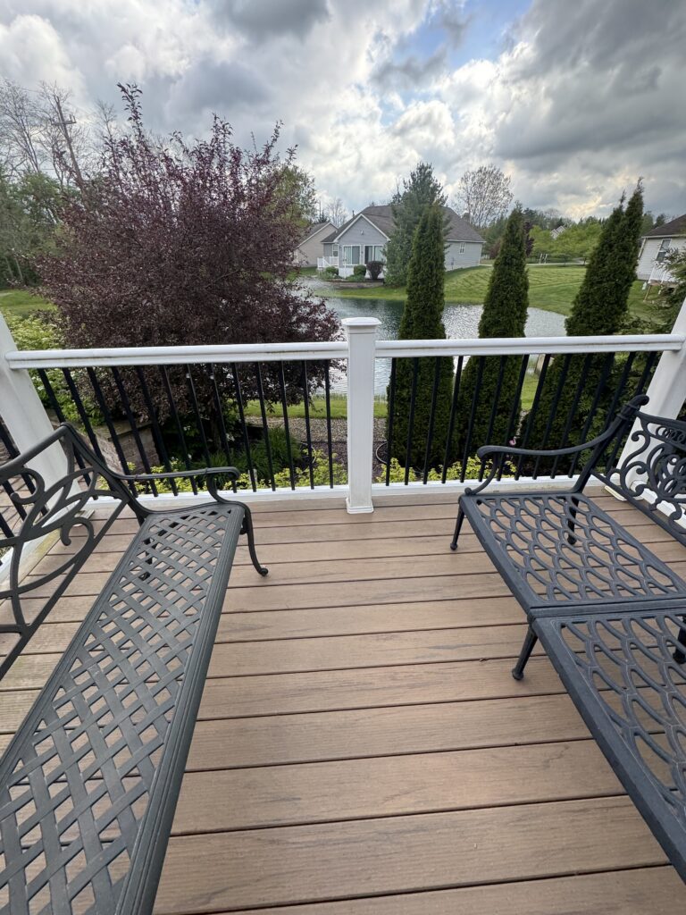 Wooden deck with black metal chairs along a railing overlooking a pond and suburban yards under cloudy skies.