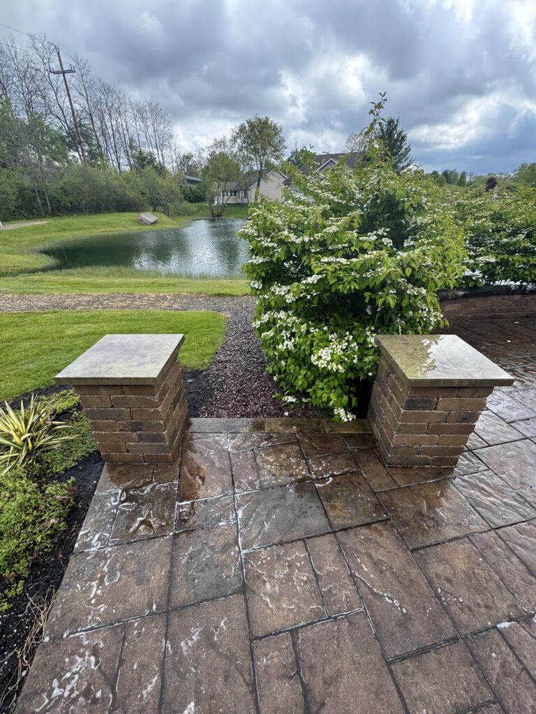 Stone-paved patio with two brick pillars and a white-flowering shrub; a pond, lawn, and house visible beyond under cloudy skies.