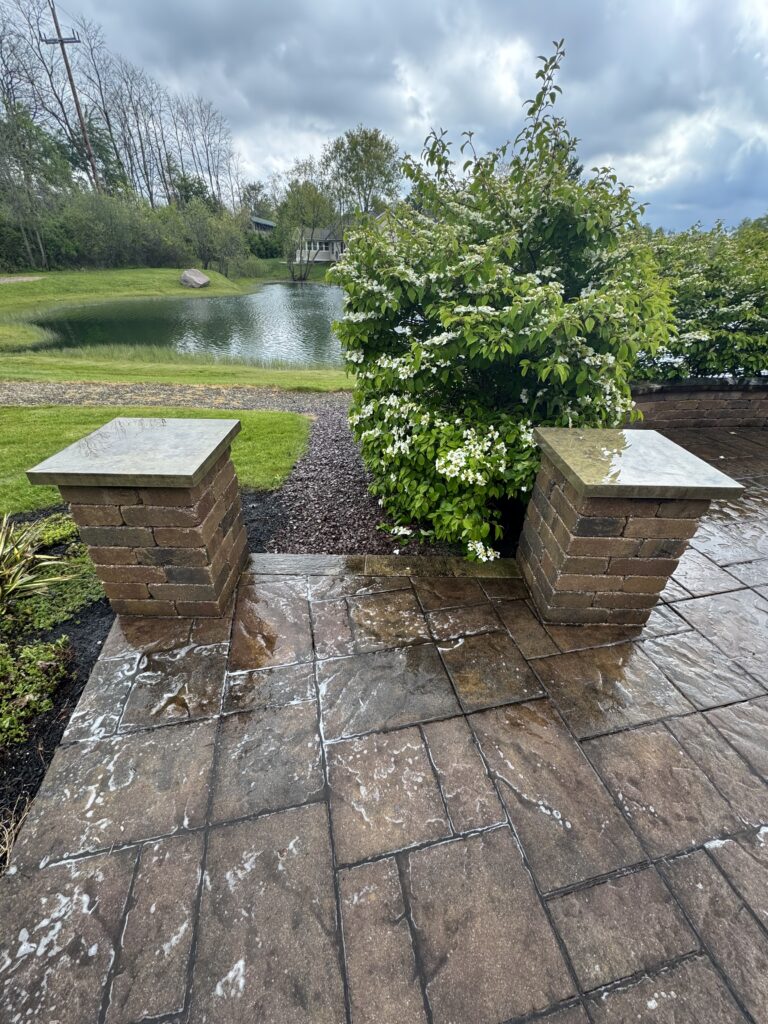 Stone patio with two brick pedestals framing a flowering white shrub; pond and cloudy sky in the background,