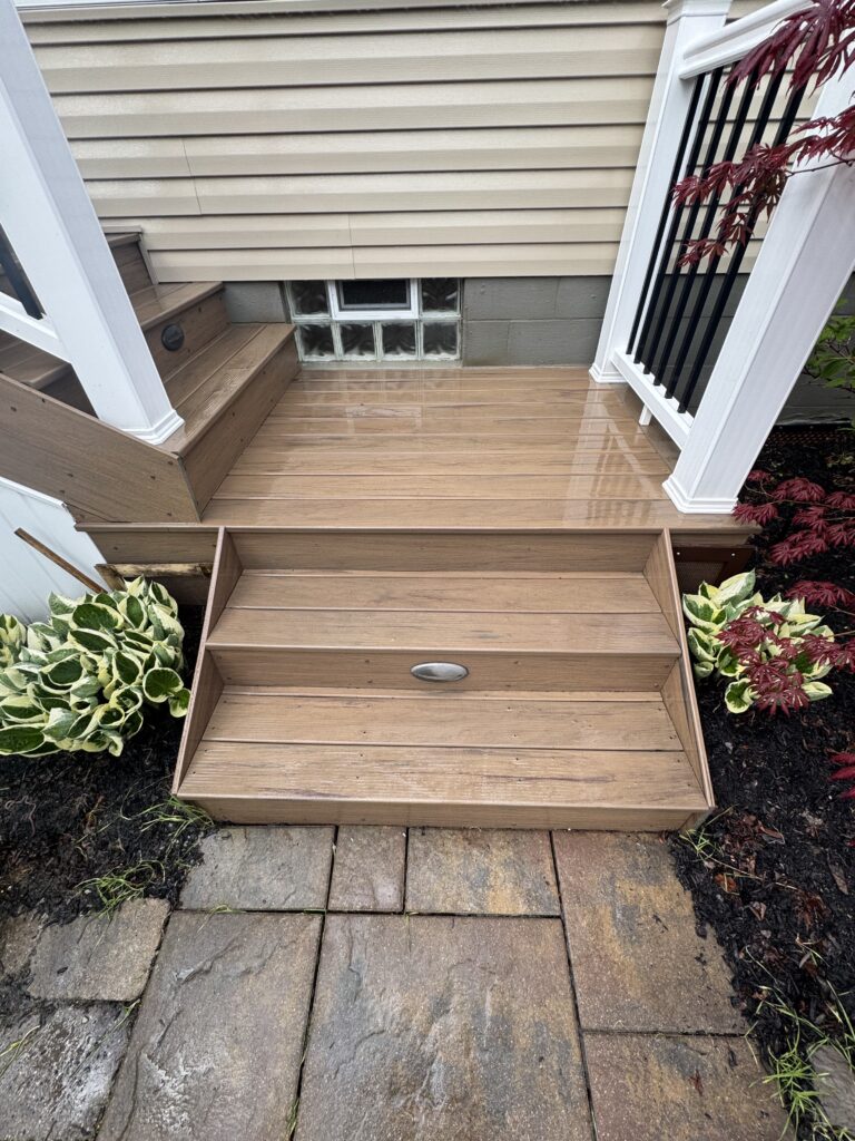 Wood deck stairs with a landing beside beige siding, a glass-block basement window, white railing with black balusters, and a red-leaf shrub nearby.