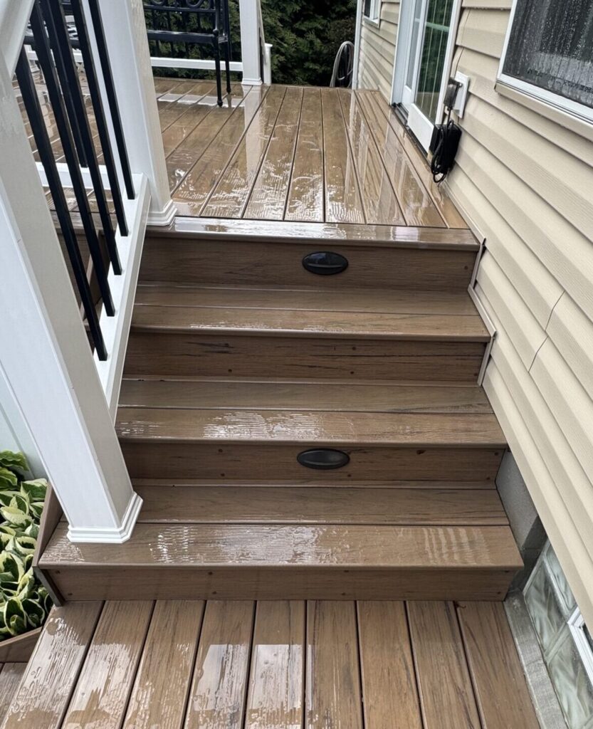 Wooden outdoor deck stairs after rain, with white posts and black balusters, beige siding, and a windowed wall beside a potted plant in the corner.