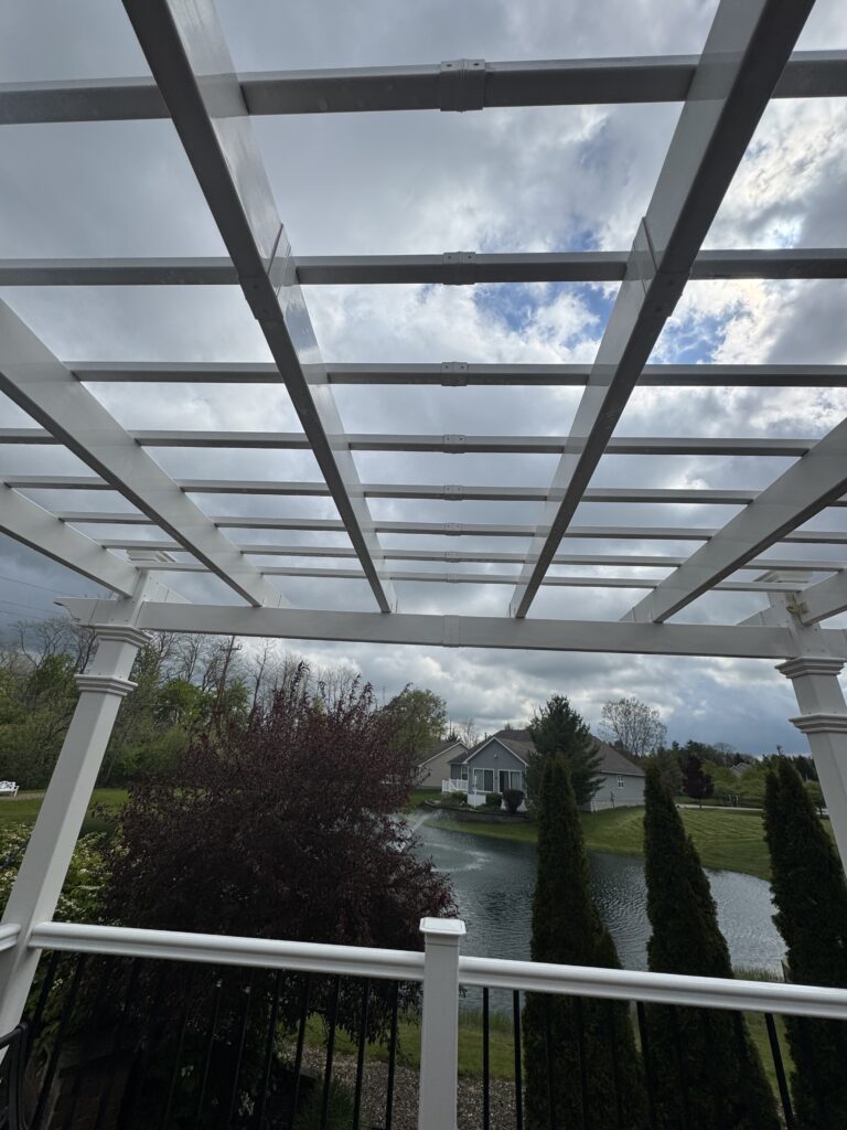 White wooden pergola framework extending over a patio, with a pond and suburban houses in the distance beneath a cloudy sky nearby trees.4