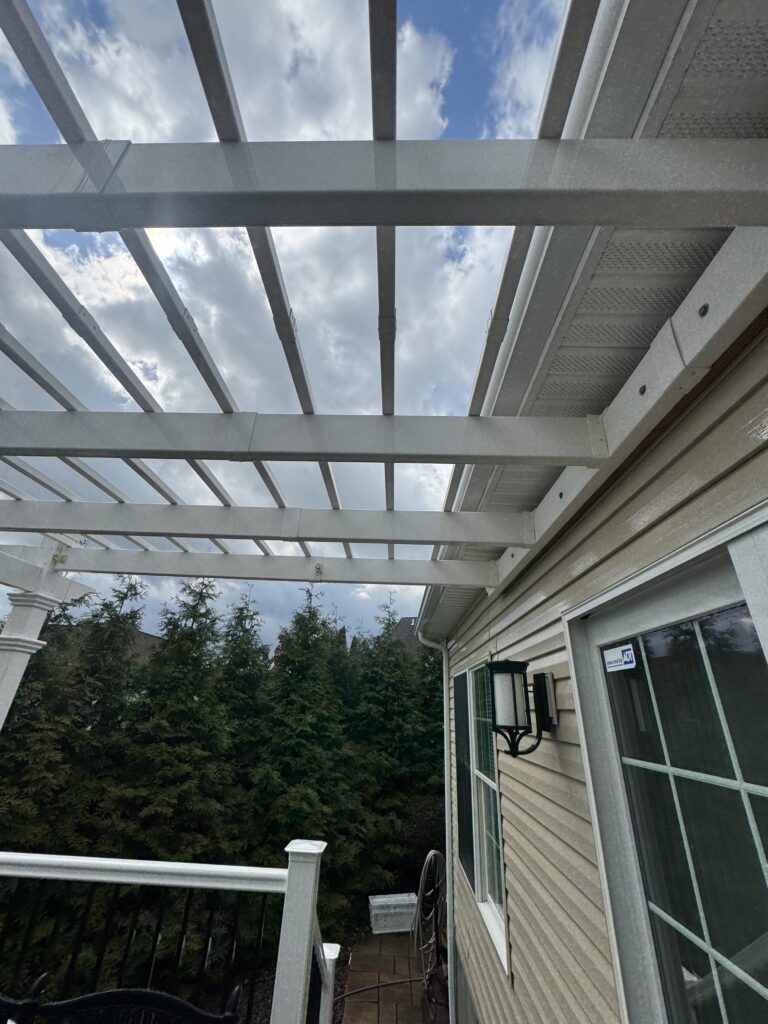 Grey metal pergola overhead with evenly spaced beams, above a stone patio beside a beige house, blue sky and clouds visible in the background
