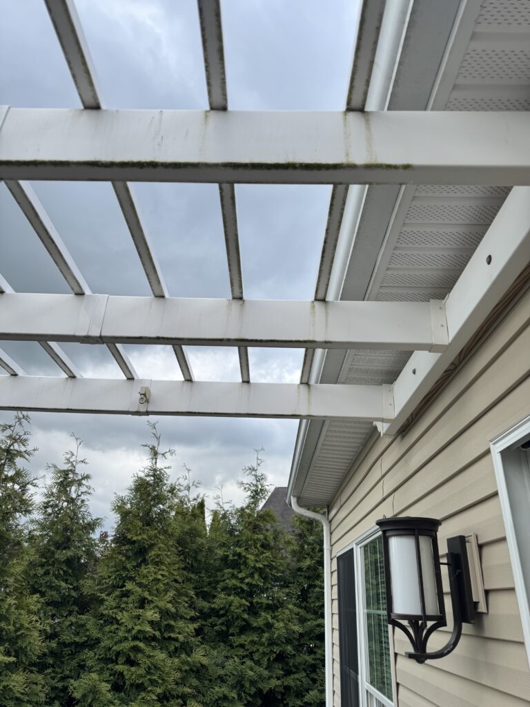 White metal pergola over a beige house exterior with moss on the beams, a black wall lantern near a window, and a cloudy sky behind.
