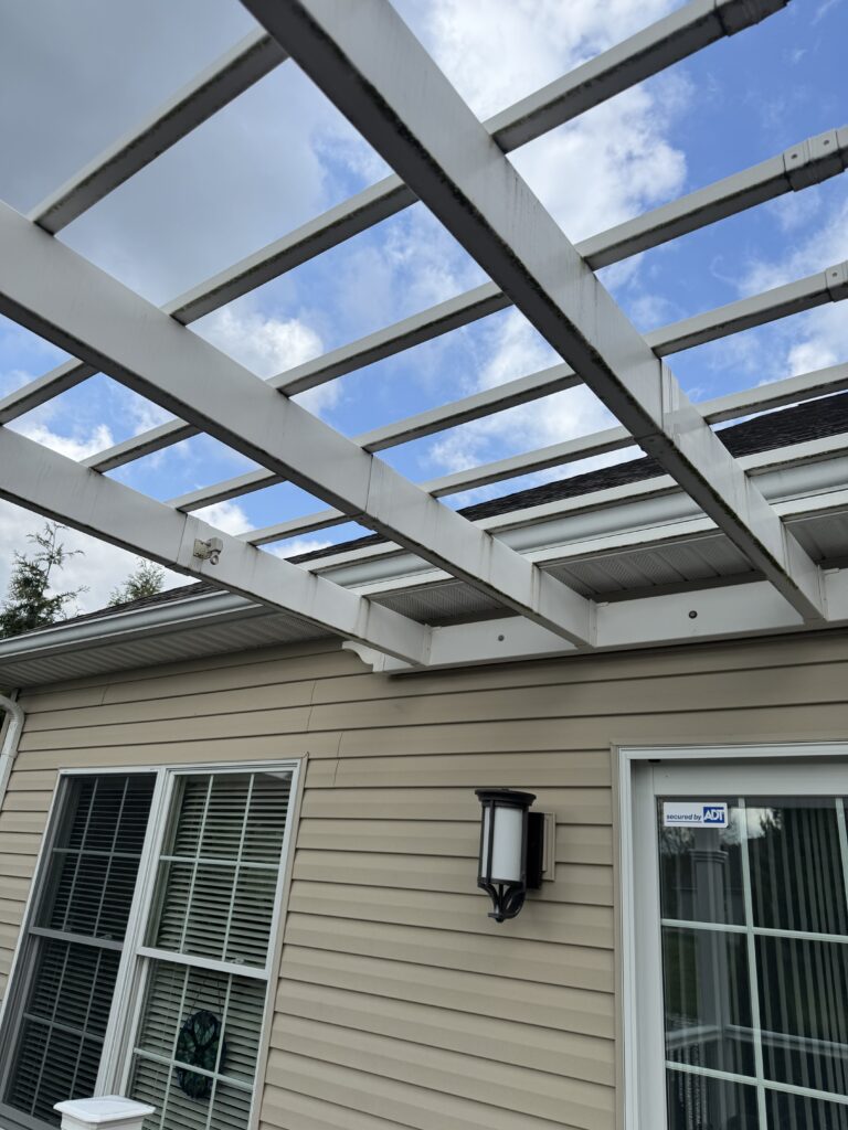 Backyard exterior: beige siding with a white wooden pergola over a patio, blue sky visible, and a wall lantern near a door with an ADT sticker