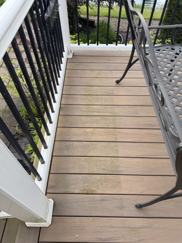 Wooden balcony deck with brown planks, black vertical railing, and a gray metal chair on the right; garden beyond.