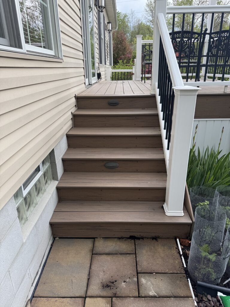 Exterior stairway: brown wooden steps leading up to a wooden deck with white rail posts and black balusters beside a beige siding house.
